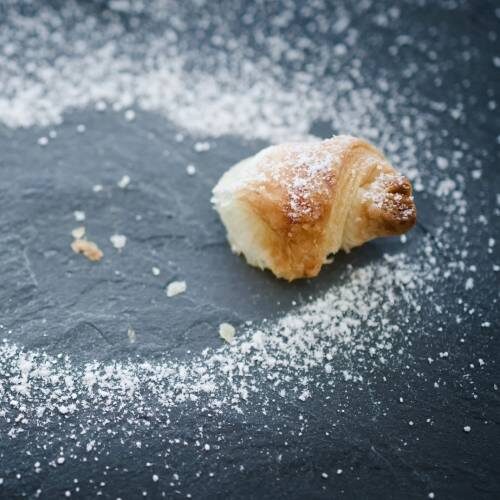 A single croissant piece surrounded by scattered powdered sugar on a dark surface.